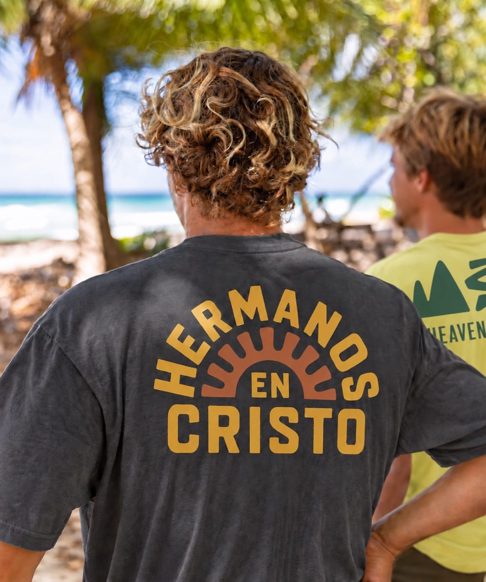 Two men at the beach wearing FIVE2 Christian graphic tees: charcoal Hermanos en Cristo and yellow Heaven on Earth.