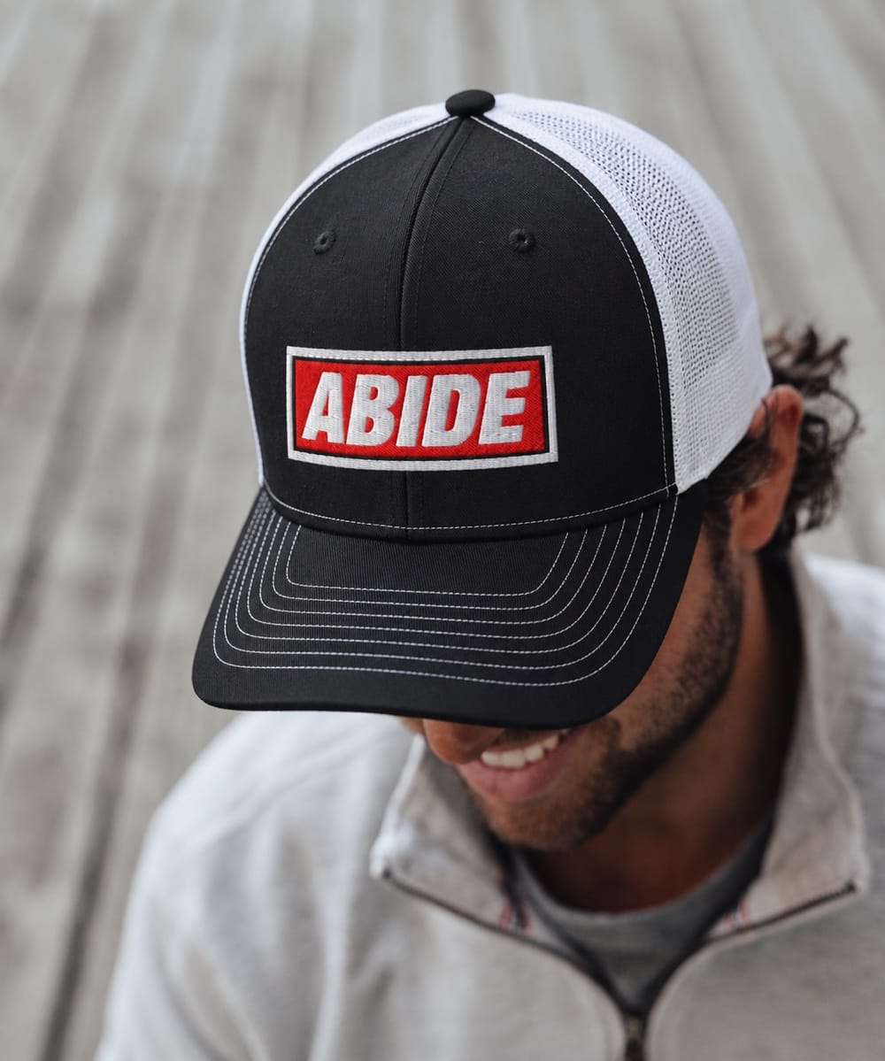 Man wearing a black and white FIVE2 Abide Christian trucker hat with a red embroidered patch.