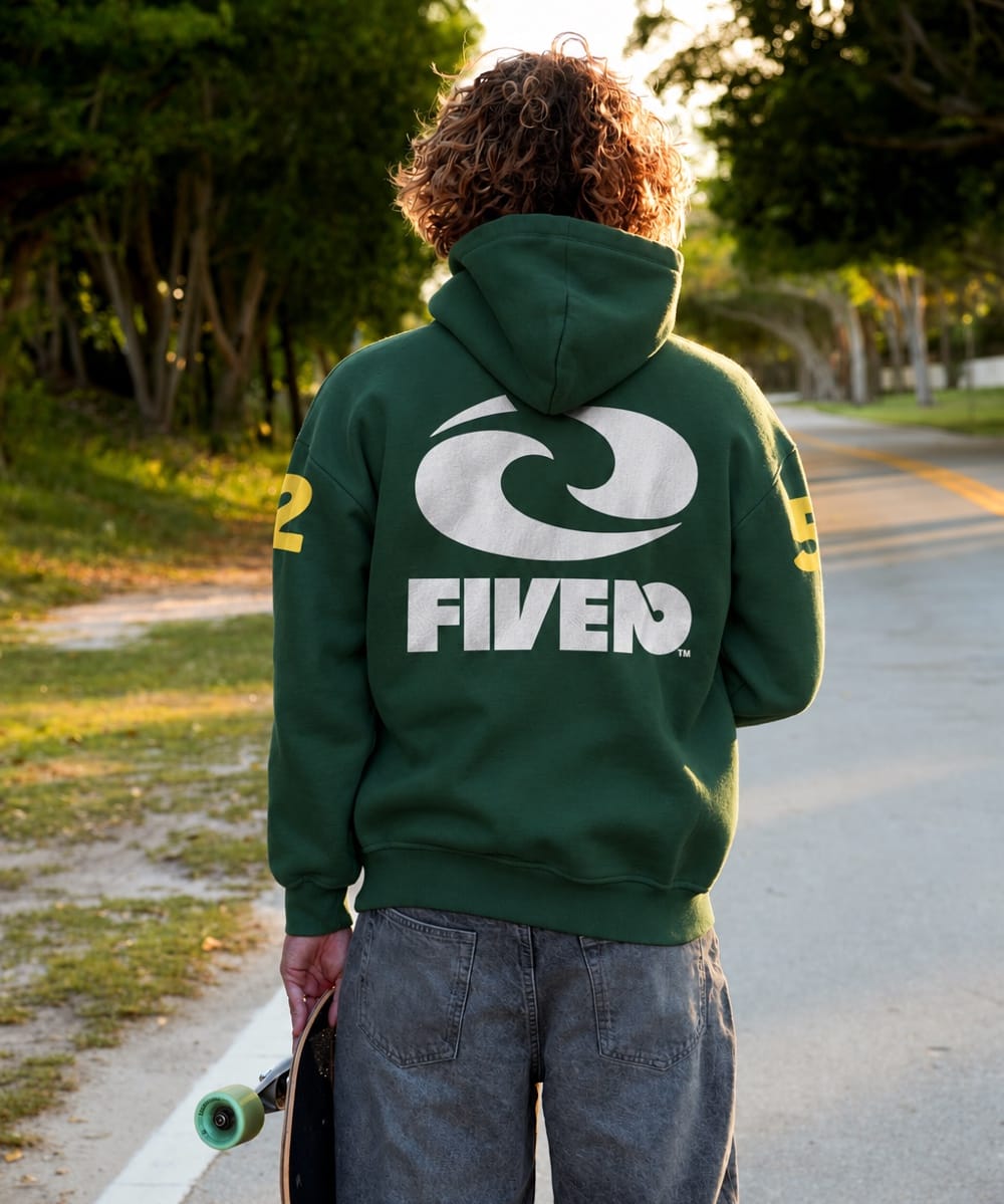 Back view of a man holding a skateboard in a forest-green FIVE2 Christian zip-up hoodie with a white wave logo and yellow sleeve graphics.